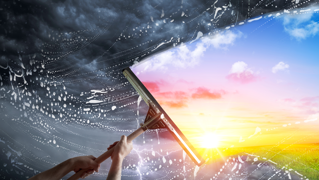 Image of a window being cleaned. It is bright on one side with a rainbow and dull and cloudy on the other side.